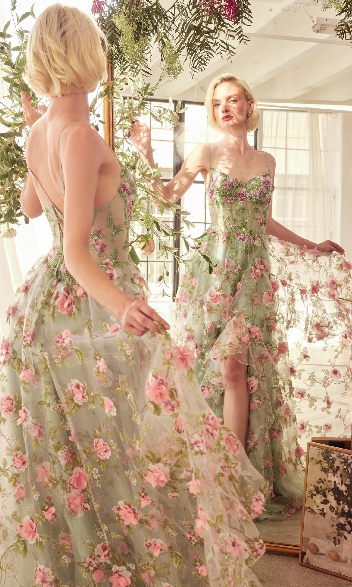 A woman in a sheer corset Sage Long Floral Prom Dress (Andrea & Leo A1420) stands before a mirror, admiring her reflection in a sunlit room filled with plants and a framed picture on the floor.